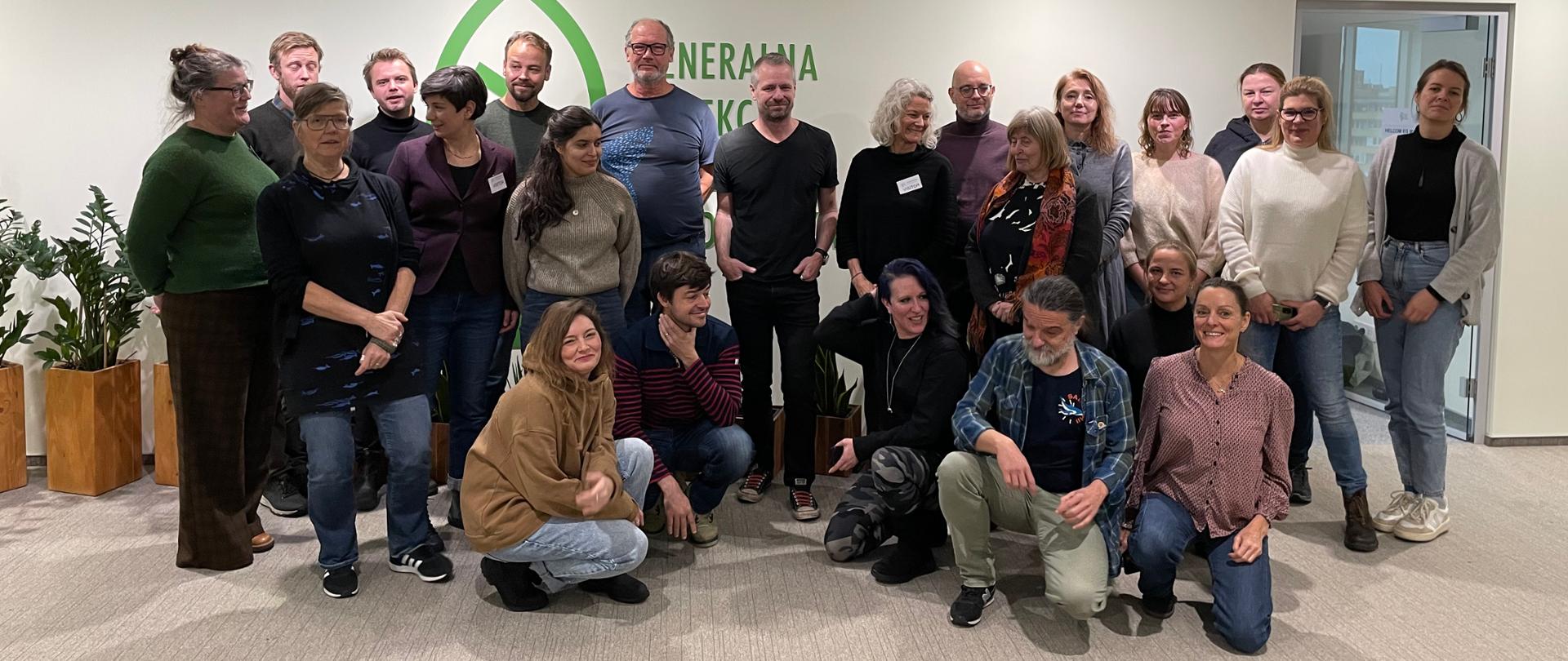 Na zdjęciu znajduje się grupa około osób dorosłych pozujących do wspólnej fotografii w pomieszczeniu biurowym. Osoby ubrane są w różnorodny sposób w stroje codzienne i casualowe (swetry, koszule, jeansy)Większość osób stoi, ale kilka jest w pozycji kucającej. Tło: ściana za nimi jest jasna, w kolorze złamanej bieli. Na ścianie znajduje się duże, zielone logo oraz napis (częściowo zasłonięty) Generalna Dyrekcja Ochrony Środowiska. 