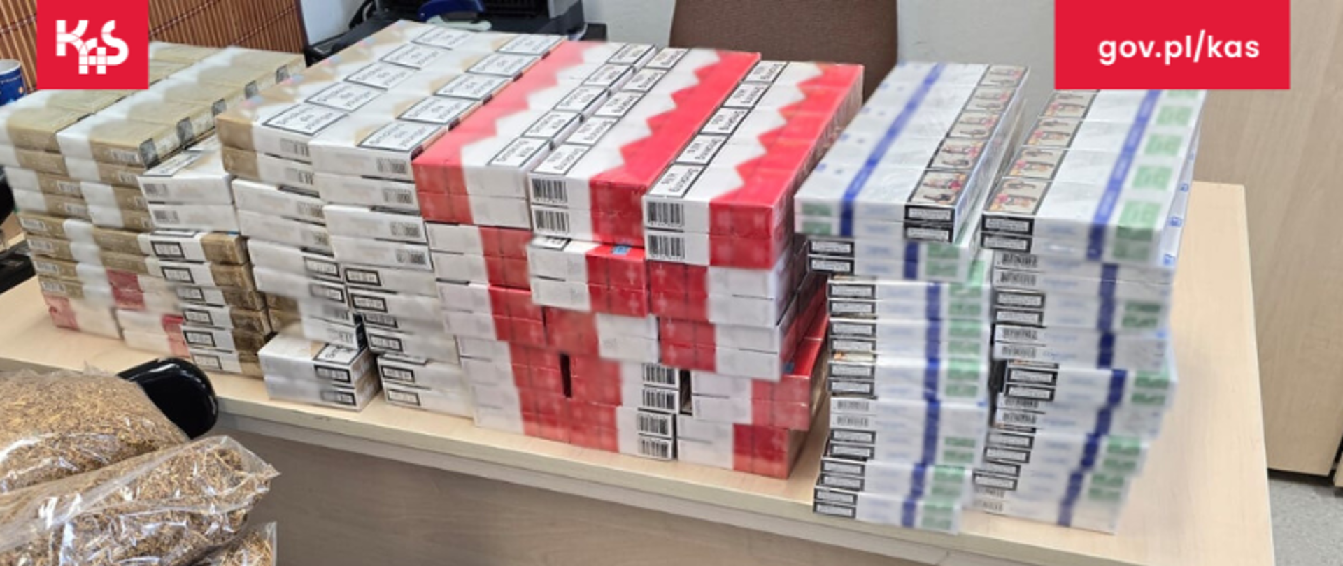 Paczki nielegalnych papierosów ułożone na biurku. Przed biurkiem paczki z nielegalnym krojonym tytoniem.