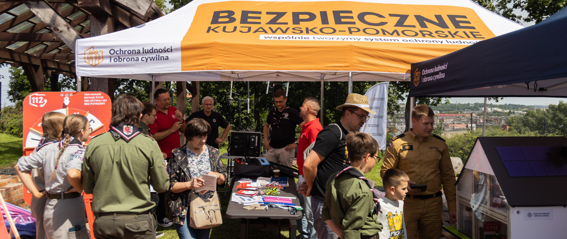 Przy namiocie zgromadzili się mieszkańcy, harcerze i przedstawiciele służb. Na stoisku znajdują się makiety oraz materiały edukacyjne. Na namiocie widoczny jest napis „Bezpieczne Kujawsko-Pomorskie”.