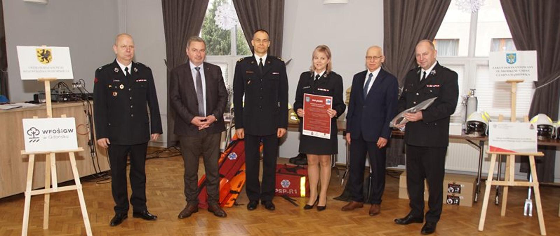 Zdjęcie przedstawia 5 mężczyzn, 3 w umundurowaniu galowym strażackim oraz 2 mężczyzn w garniturach, a także 1 kobietę w umundurowaniu galowym ustawionych obok sztalug z tablicami informacyjni oraz sprzętem do ratownictwa medycznego