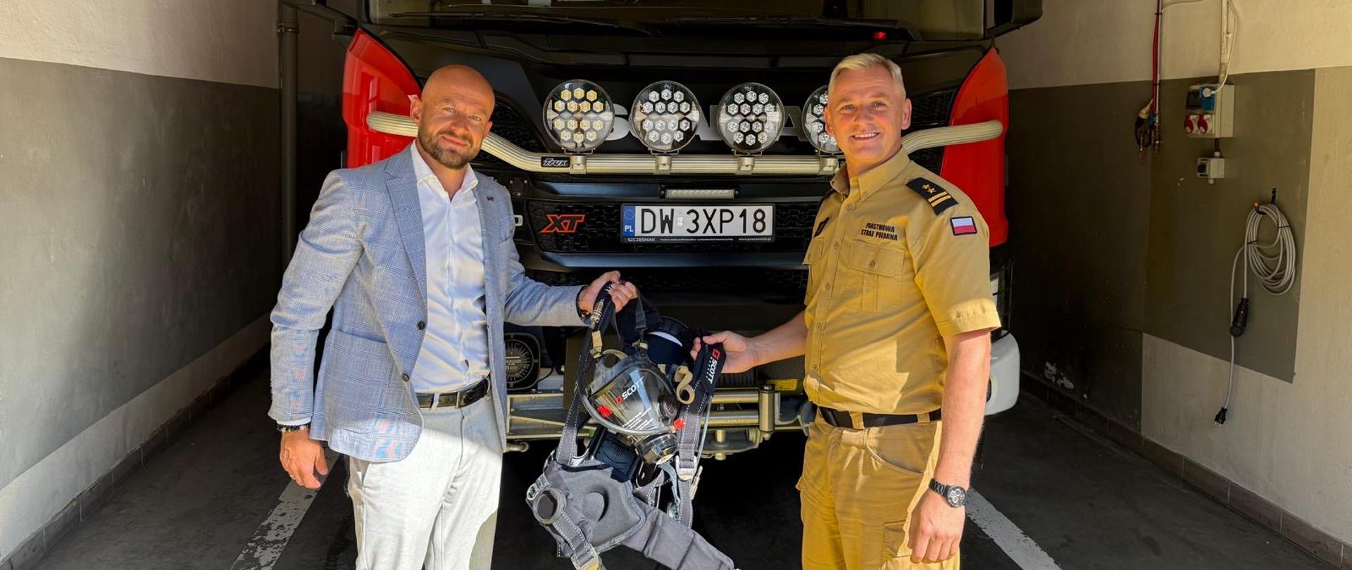 Na zdjęciu uwieczniono moment oficjalnego przekazania sprzętu strażackiego w garażu remizy. Mężczyzna w garniturze oraz funkcjonariusz Państwowej Straży Pożarnej w mundurze służbowym razem trzymają nowoczesny aparat powietrzny z maską oddechową. W tle za nimi stoi ciężki wóz strażacki marki Scania. Zdjęcie ma charakter symbolicznego przekazania nowego wyposażenia dla jednostki.