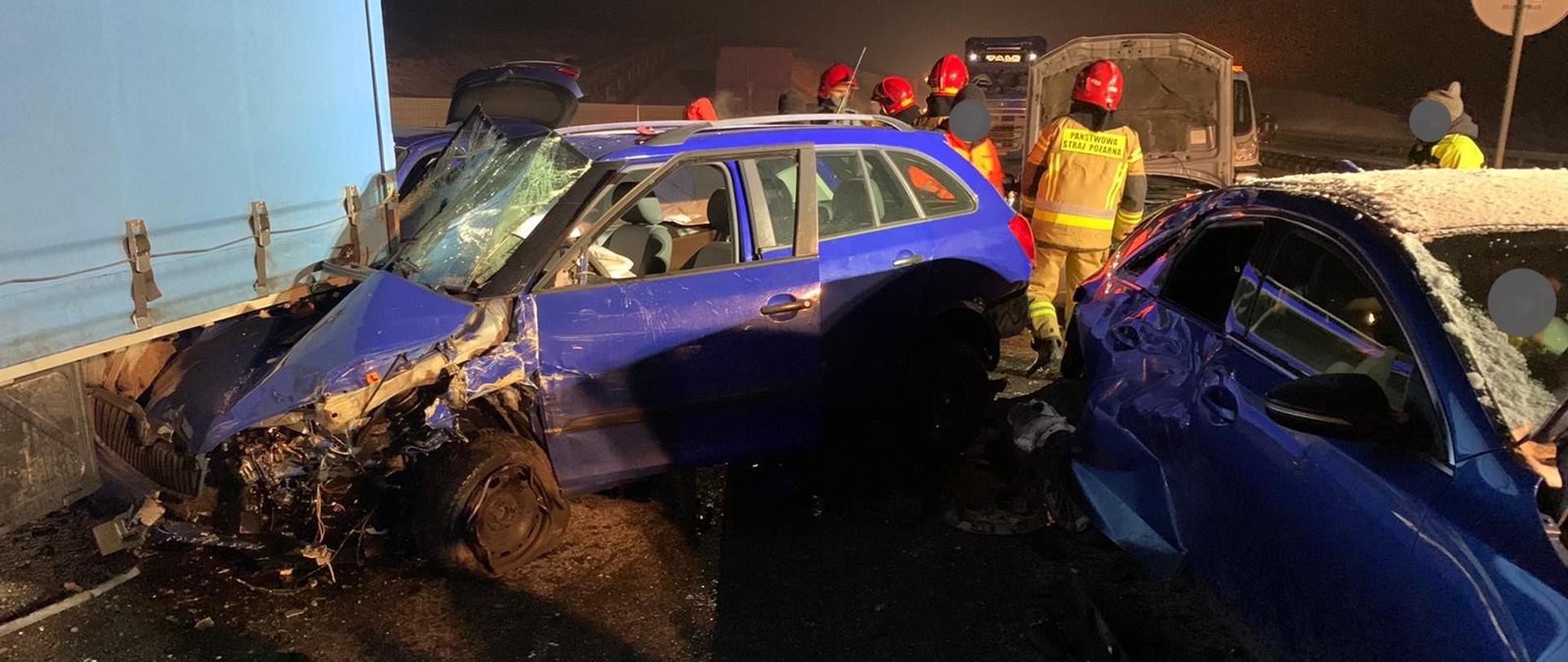 Na zdjęciu widoczne są dwa uszkodzone samochody w kolorze niebieskim. Z lewej strony widoczna tylna część samochodu dostawczego pod którego tył wbity jest przód samochodu osobowego. Na drugim planie widoczni są strażacy oraz kolejne uszkodzone pojazdy.
