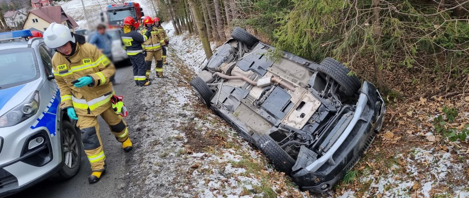 Samochód osobowy na dachu poza drogą. Obok strażacy oraz samochody służb.