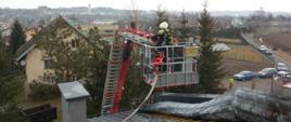 Na zdjęciu strażak w koszu podnośnika hydraulicznego monitorujący temperaturę poszycia dachu w celu lokalizacji zarzewi ognia. dach budynku kryty blachodachówką koloru antracyt ze zdjętym jednym arkuszem blachy, pod którą rozwijał się pożar.