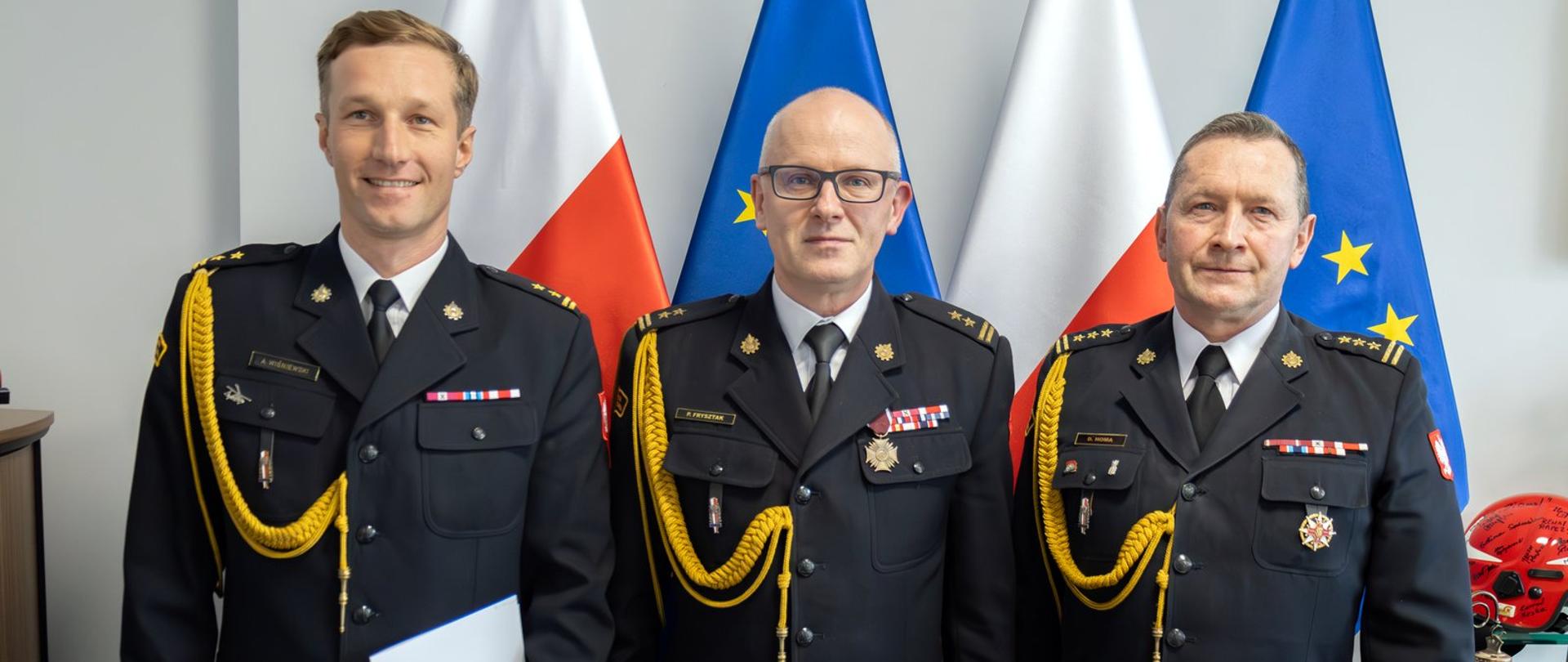 Zdjęcie zrobione wewnątrz pomieszczenia. Na fotografii stoi trzech oficerów Państwowej Straży Pożarnej w mundurach galowych. Za nimi ustawione są flagi RP i UE.