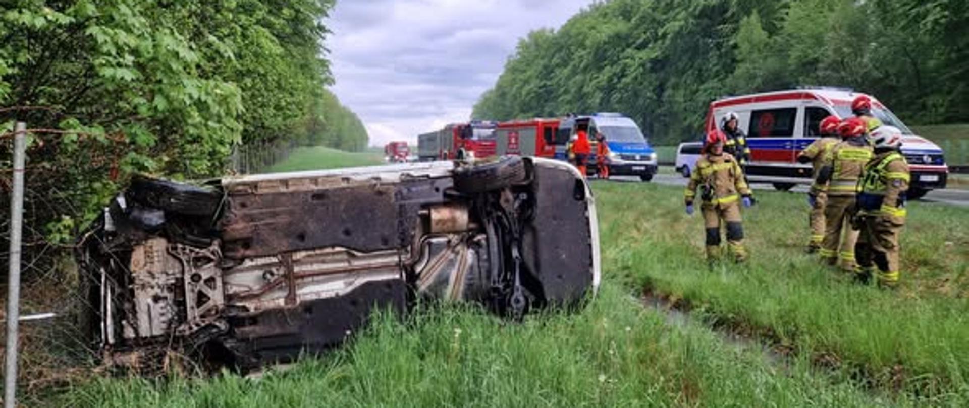 Na zdjęciu znajduje się wrak samochodu osobowego leżący na boku. W tle stoją samochody strażackie, radiowóz i karetka pogotowia.