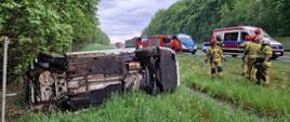 Na zdjęciu znajduje się wrak samochodu osobowego leżący na boku. W tle stoją samochody strażackie, radiowóz i karetka pogotowia.