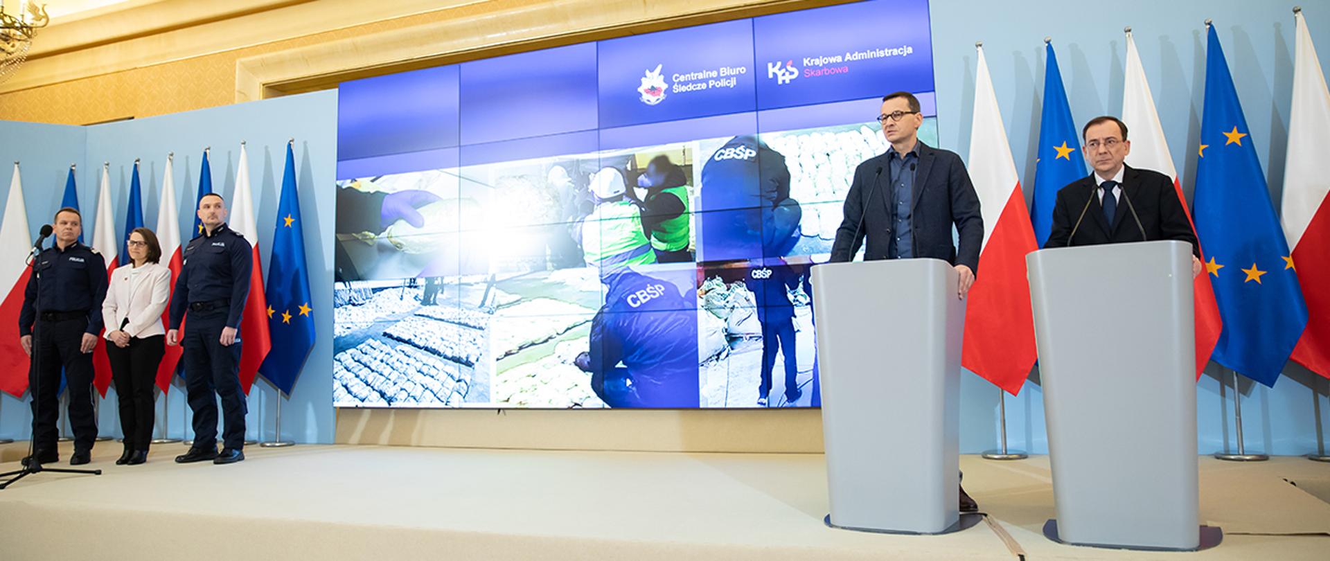 Na zdjęciu widać stojących na scenie przy mównicach w trakcie konferencji prasowej premiera Mateusza Morawieckiego i ministra Mariusza Kamińskiego. Na scenie znajdują się również szef Krajowej Administracji Skarbowej Magdalena Rzeczkowska, nadinsp. Dariusz Augustyniak – Zastępca Komendanta Głównego Policji i insp. Paweł Półtorzycki – Komendant CBŚP. Premier zabiera głos.