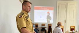 Strażak, w mundurze służbowym stoi przed stołem i prezentuje slajdy. Przed nim widać dwoje innych uczestników spotkania. W tle, na ścianie wyświetlany slajd ze strażakiem.