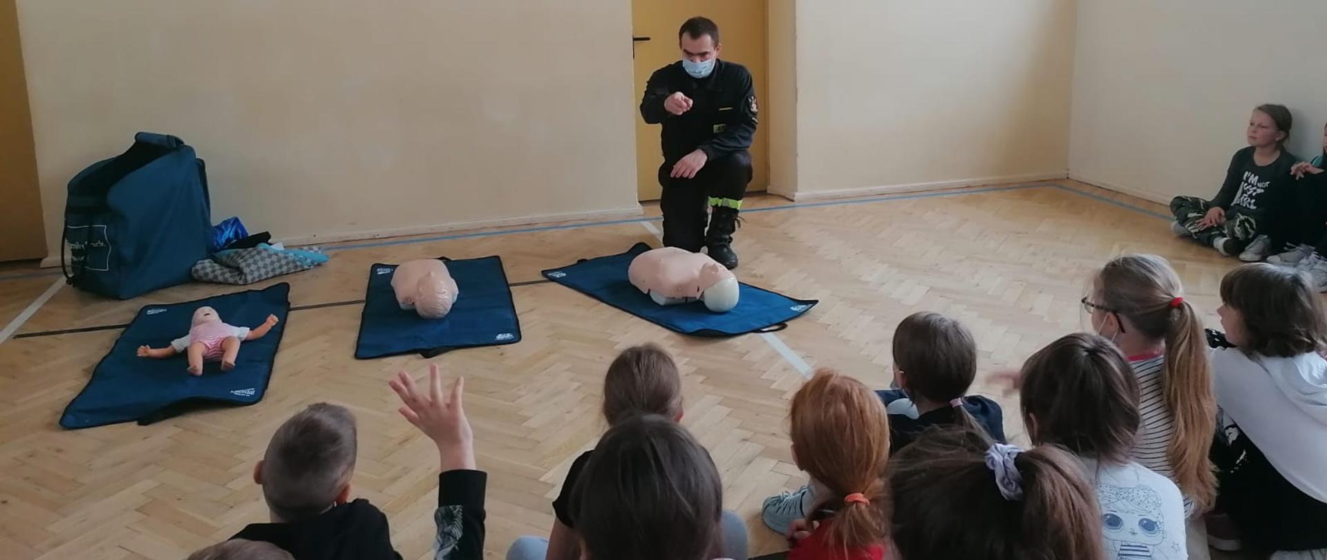 Strażak klęczy na jednym kolanie przy rozłożonych na matach trzech manekinach. Przed nim siedzą dzieci. Jando z nich podnosi do góry rękę.