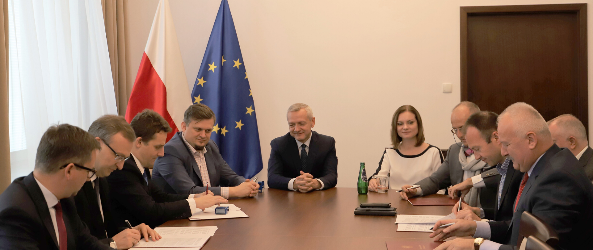 Przy owalnym stole siedzi kilka osób. Część z nich podpisuje dokumenty. Na środku siedzi minister Marek Zagórski. Za nim stoją flagi polska i unijna.