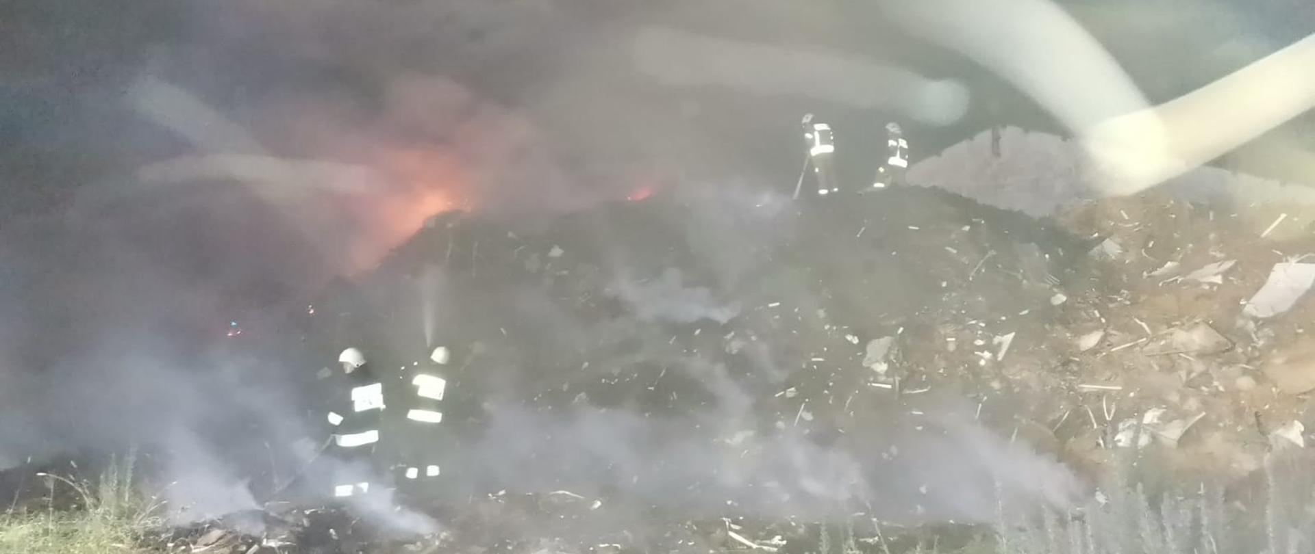 Fotografia przedstawia ujęcie hałdy pozostałości po meblach o wysokości około 5 metrów. Jest noc, widoczne płomienie na szczycie hałdy. U dołu hałdy i na szczycie hałdy stoją roty gaśnicze – po dwóch strażaków w ubraniach specjalnych i w hełmach – podają prądy wody w natarciu na pożar. Panuje zadymienie, w tle za hałdą widoczny szczyt budynku magazynowego.