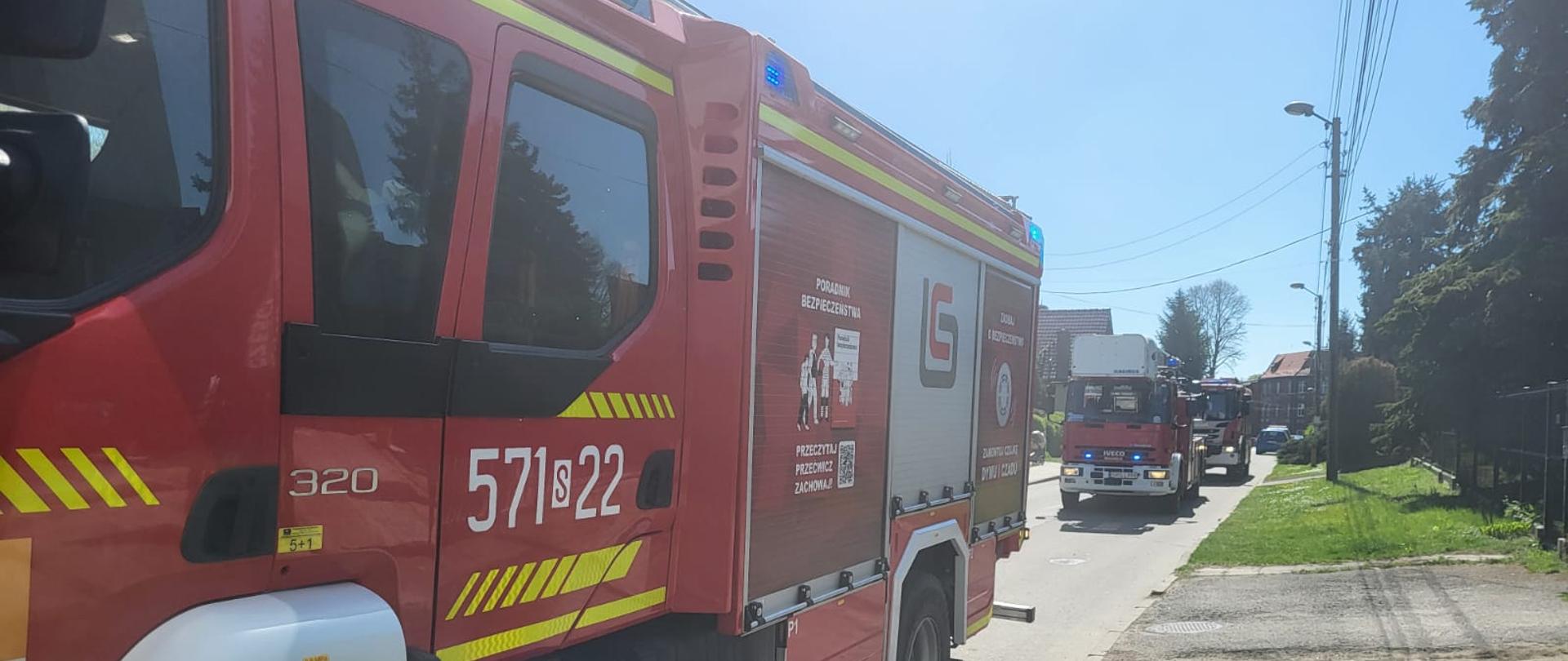 Dwa czerwone samochody straży pożarnej stojące na wąskiej ulicy w zabudowie jednorodzinnej; na pierwszym planie widoczny bok wozu ratowniczo-gaśniczego, w tle kolejny pojazd z włączonymi sygnałami świetlnymi.