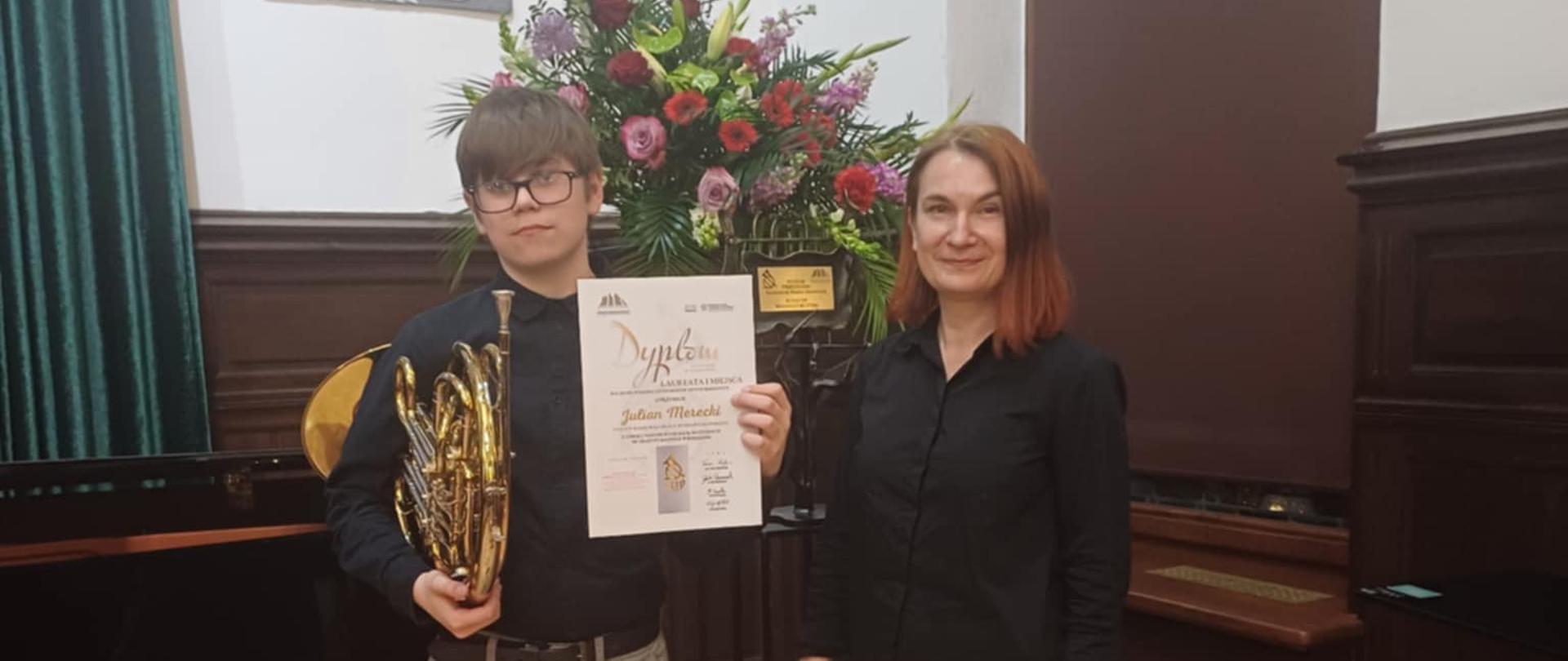 Uczeń z waltornią i dyplomem, z boku stoi nauczycielka, w tle kwiaty.