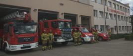 Na zdjęciu widać ustawione przed garażami samochody pożarnicze z włączonymi sygnałami świetlnymi oraz ustawionych z boku samochodów strażaków w ubraniu specjalnym.