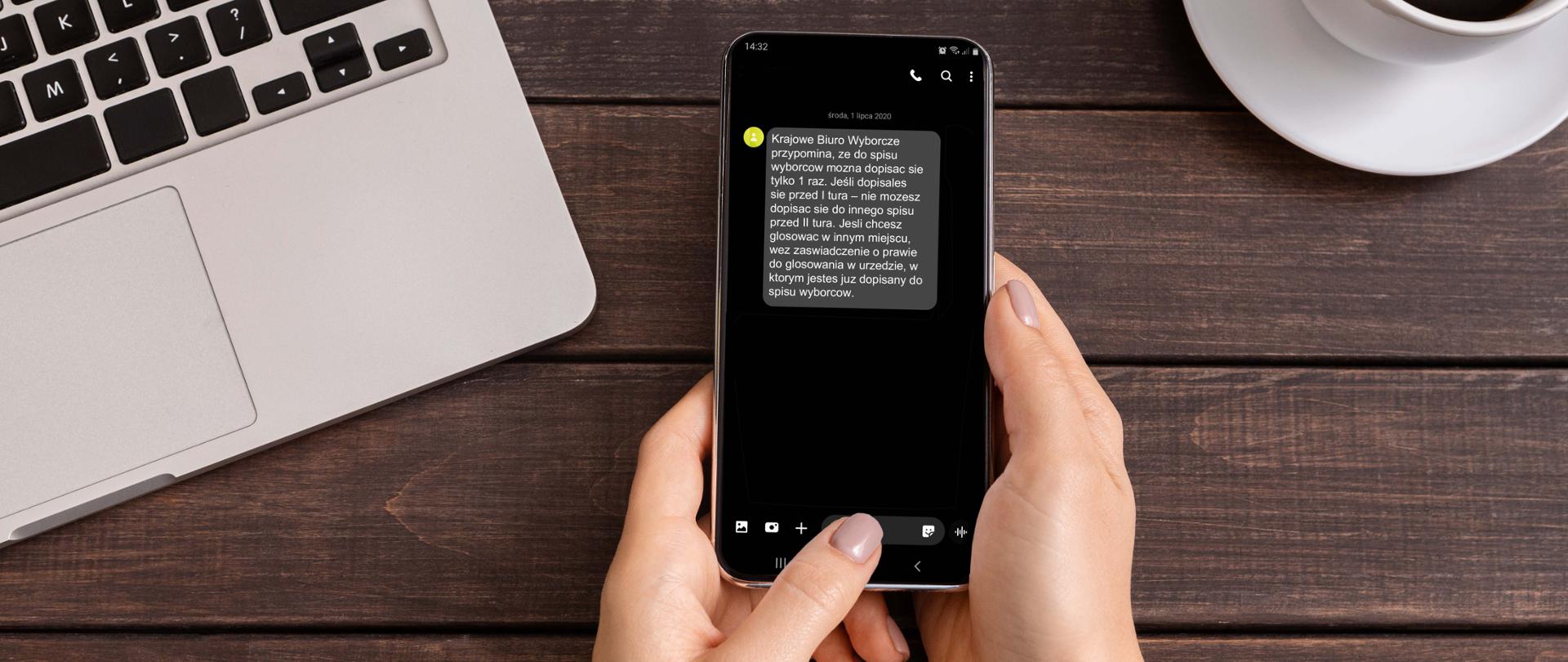 Biurko, na którym stoją laptop i filiżanka z kawą. Na pierwszym planie kobiece dłonie ze smartfonem. Na ekranie wyświetla się komunikat: Krajowe Biuro Wyborcze przypomina, że do spisu wyborców można się dopisać tylko 1 raz. Jeśli dopisałeś się przed 1 turą - nie możesz dopisać się do innego spisu przed II turą. Jeśli chcesz głosować w innym miejscu, weź zaświadczenie o prawie do głosowania w urzędzie,w którym jesteś dopisany do spisu wyborców,