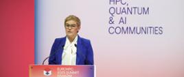 Wiceminister Mrówczyńska stoi za mównicą z napisem Euro HPC Summit 2025 Kraków i mówi do dwóch mikrofonów, za nią na ścianie napis Connecting HPC, Quantum & AI Communities.