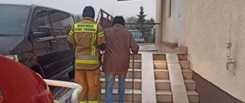 Strażak w piaskowym ubraniu prowadzi pod rękę starszą osobę z laską po stromych zaśnieżonych schodach do budynku