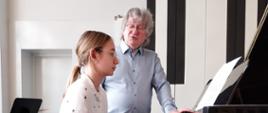 Dziewczyna siedzi przy fortepianie, obok za nią stoi mężczyzna.