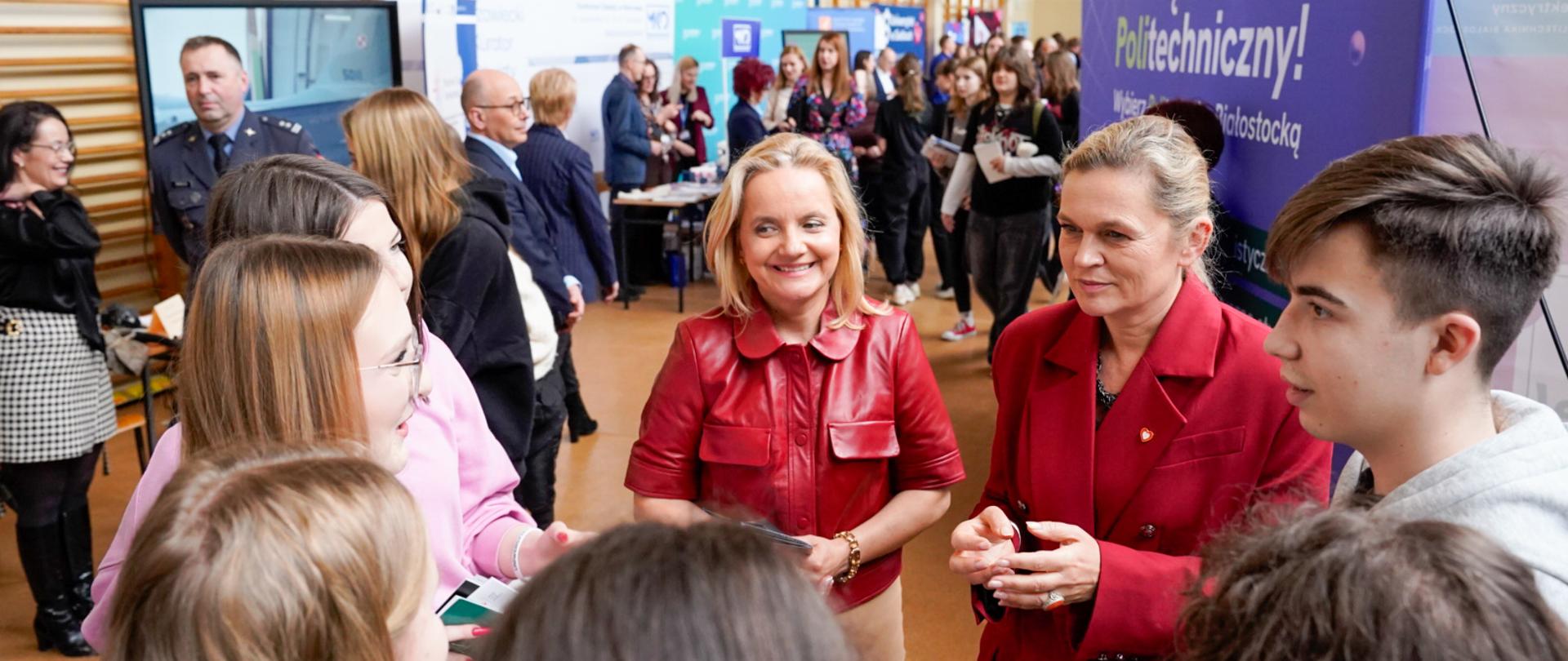 Barbara Nowacka rozmawia z grupą uczniów w hali sportowej. Minister intensywnie gesticuluje, tłumacząc coś młodzieży. W tle widoczne są stoiska m.in. Politechniki Białostockiej.