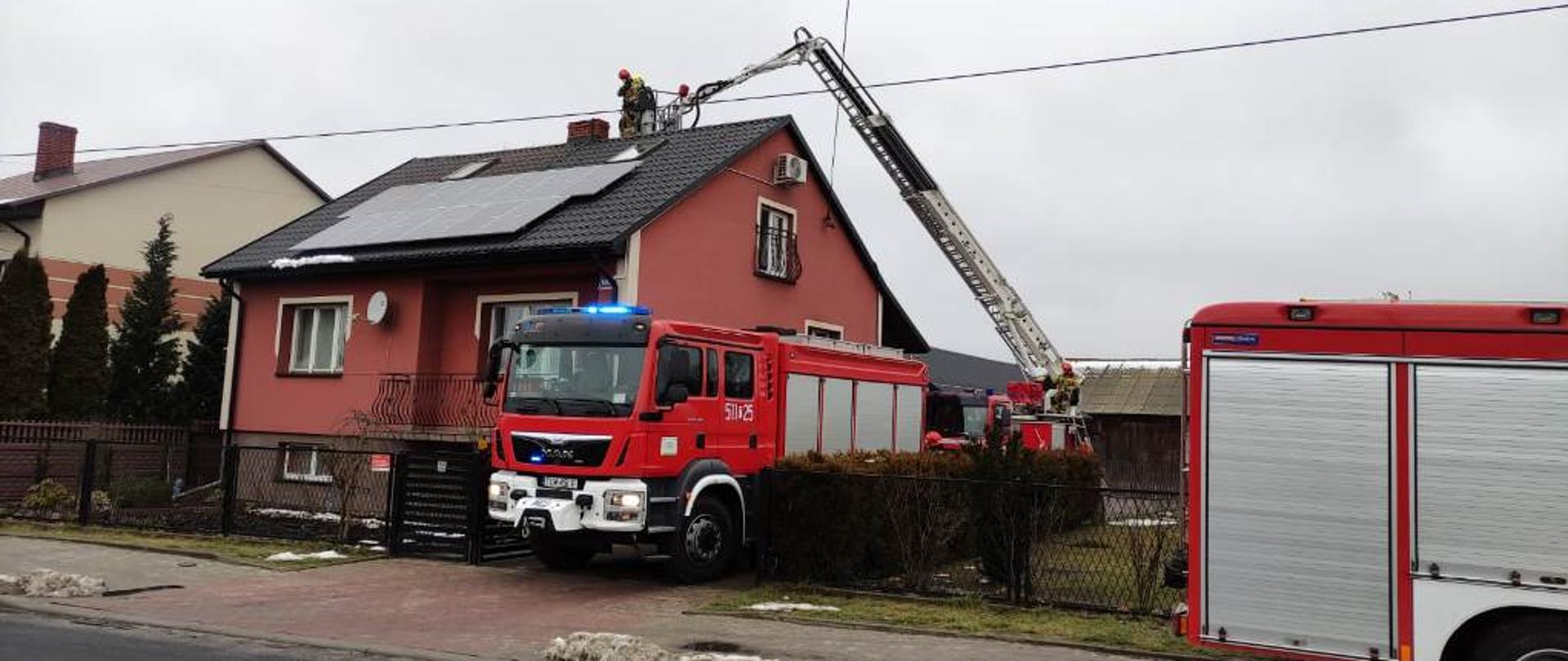 Na pierwszym planie pojazdy ratowniczo-gaśnicze , nieco dalej pojazd specjalny podnośnik hydrauliczny, w koszu podnośnika strażak prowadzący działania gaśnicze podczas pożaru sadzy w kominie budynku mieszkalnego znajdującego się na drugim planie.