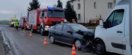 Na zdjęciu widać kolizję dwóch samochodów. Zielony samochód osobowy przodem uderzył w białego busa. Pojazdy stoją na asfaltowej jezdni. Za nimi stoją strażacy w czerwonych i białych hełmach. Dalej zaparkowane są dwa czerwone samochody strażackie, radiowóz policyjny oraz żółta karetka pogotowia. Obok pojazdów straży pożarnej stoi pani policjantka w żółtej odblaskowej kamizelce. Na jezdni ustawione są cztery biało-pomarańczowe pachołki drogowe. W tle widać budynek mieszkalny, drzewa oraz pole. Jest widno.