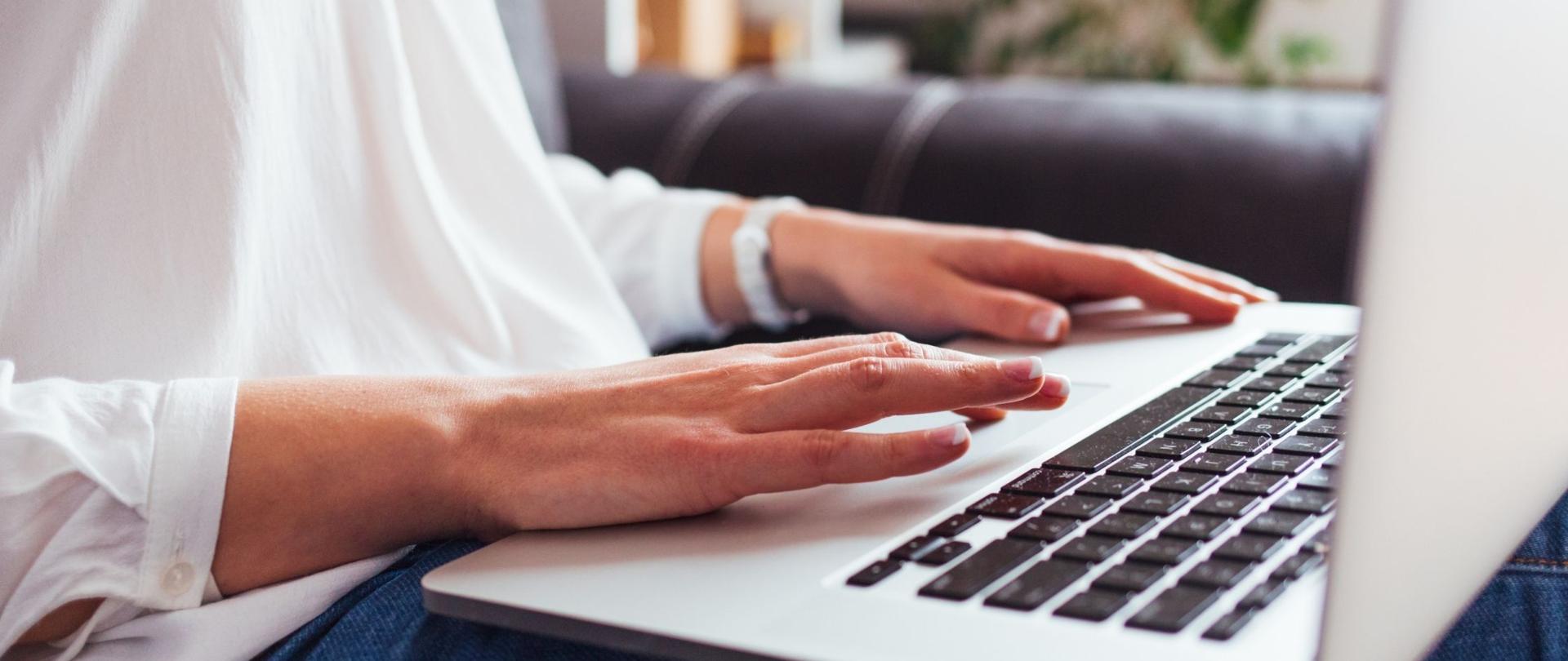 Na zdjęciu widoczna kobieta pracująca przy laptopie. Biała bluzka, ciemne spodnie. 