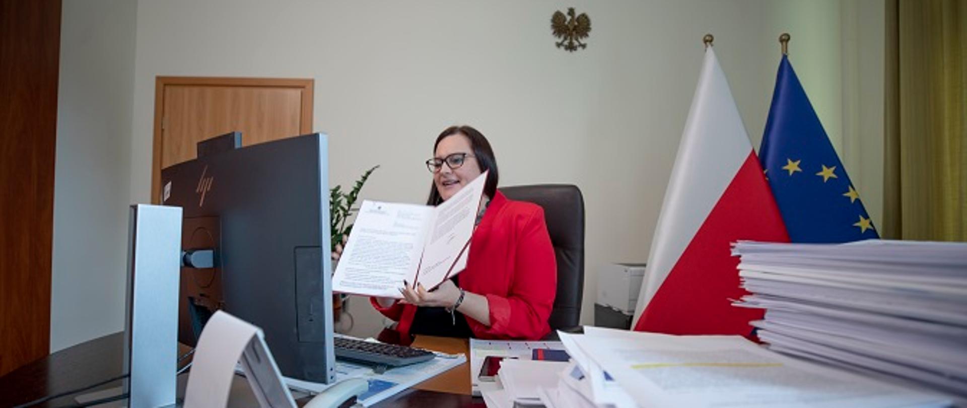 Wiceminister Małgorzata Jarosińska-Jedynak w swoim gabinecie, siedzi przy biurku i w rękach trzyma dokument - decyzję o dofinansowaniu prac nad dokumentacją potrzebnej do budowy mostu w Nowym Brzesku.