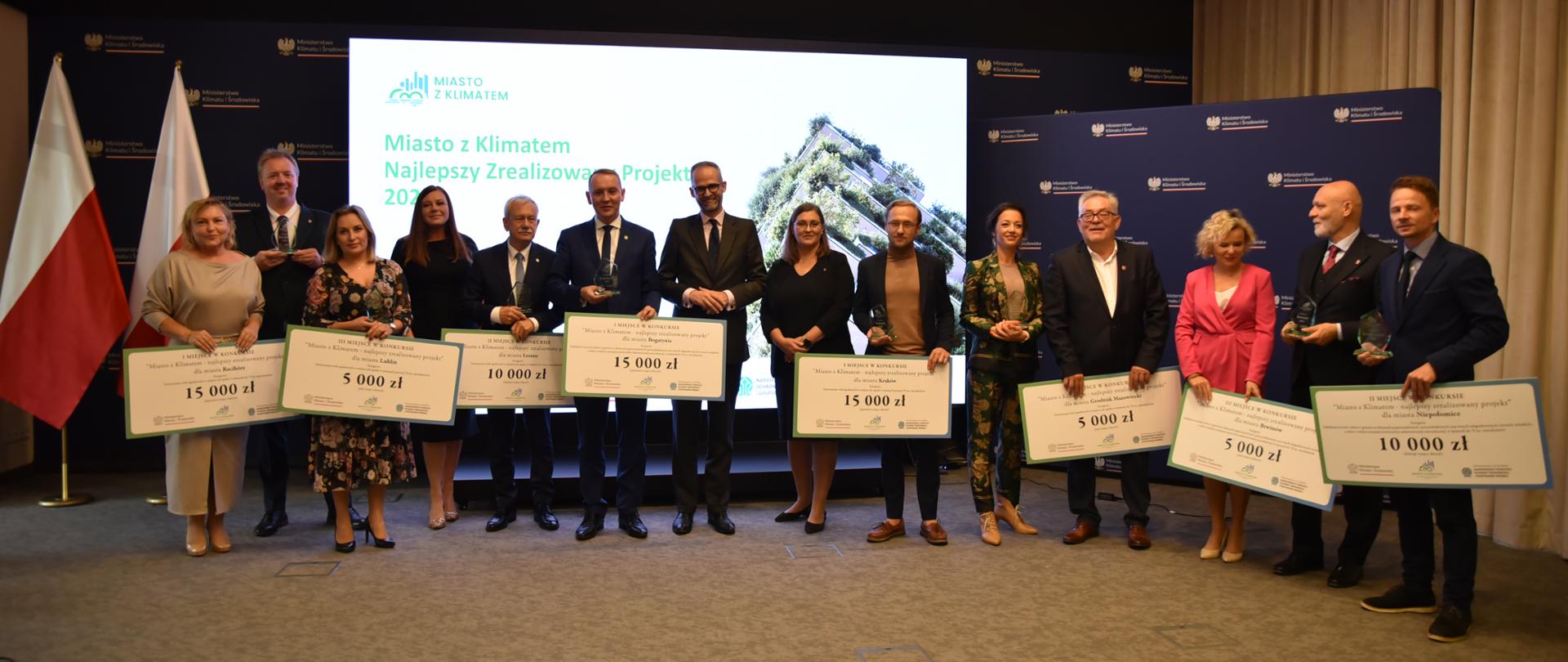 Grupowe zdjęcie wszystkich laureatów IV edycji konkursu Miasto z klimatem - najlepszy zrealizowany projekt, trzymających w rękach statuetki i symboliczne czeki