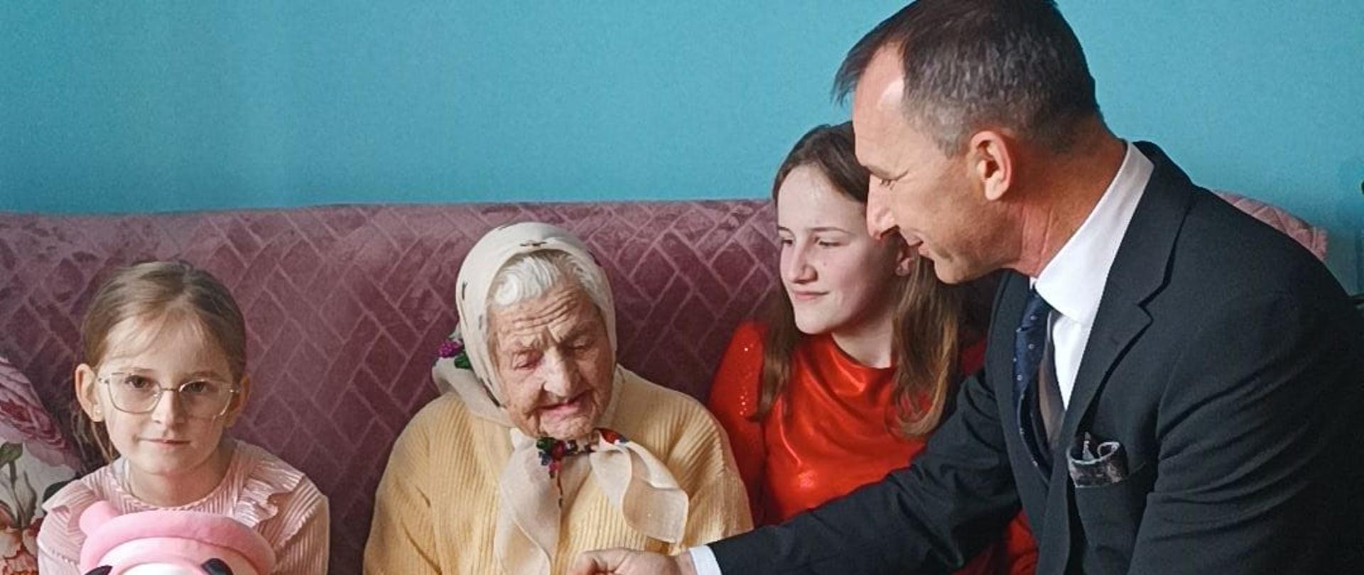 Jubilatka siedząc na sofie w towarzystwie swoich dwóch prawnuczek odbiera list gratulacyjny od Prezesa KRUS.