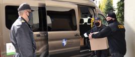 Na zdjęciu Komendant Powiatowy PSP w Ropczycach w ubraniu dowódczo-sztabowym nadzoruje przekazanie pudełek z maseczkami jednorazowymi do samochodu dostawczego. Strażak oraz dowódca JRG przenoszą pudełka z maseczkami.