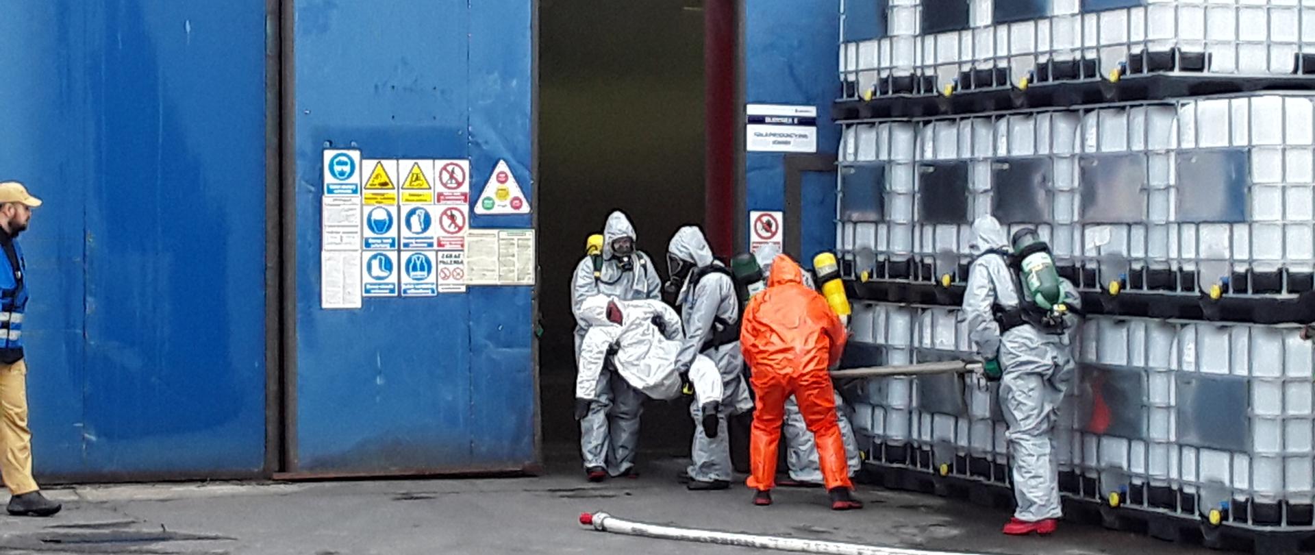 Funkcjonariusz Straży Pożarnej w srebrnych i pomarańczowych strojach ochronnych wynoszą poszkodowanego z hali.