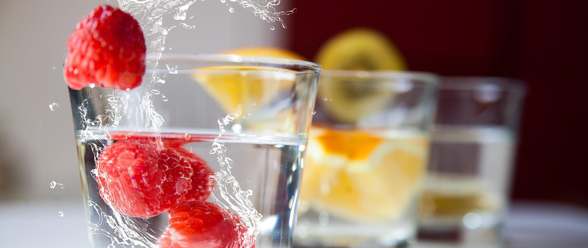 Na zdjęciu znajduje się szklanka z wodą i malinami. W tle (widok rozmazany) są dwie kolejne szklanki z wodą i owocami.