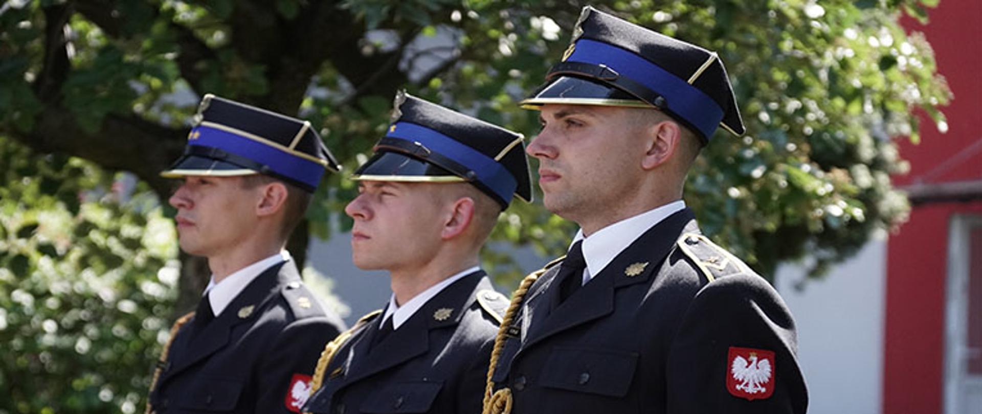 trzech strażaków ubranych w mundury galowe z rogatywką na głowie stoją w pozycji zasadniczej i patrzą przed siebie, w tle drzewa i budynek komendy