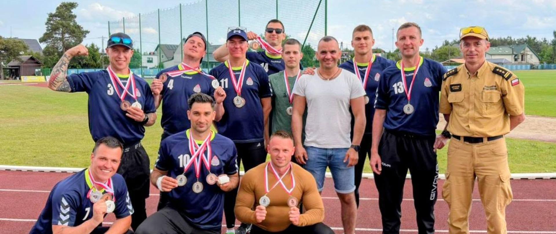 Zdjęcie przedstawia mężczyzna wchodzących w skład drużyny sportowej, ubranych w stroje spotowe, wraz z zdobytymi medalami i pucharami.