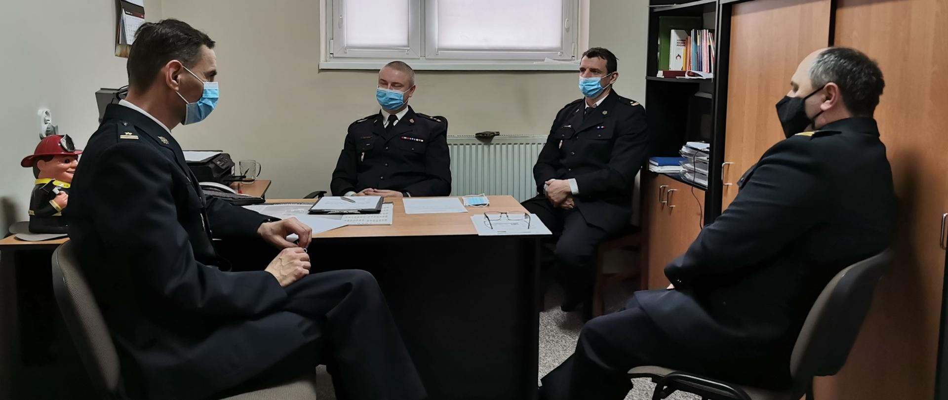 Zdjęcie przedstawia czterech strażaków w umundurowaniu wyjściowym, biorących udział w videokonferencji z Komendantem Wojewódzkim PSP w Toruniu, siedzących na krzesłach przy burku na którym znajdują się dokumenty oraz monitor. W górnej części zdjęcia widoczne jest okno zasłonięte roletą. Z prawej części zdjęcia widoczny regał z książkami oraz dekoracjami.