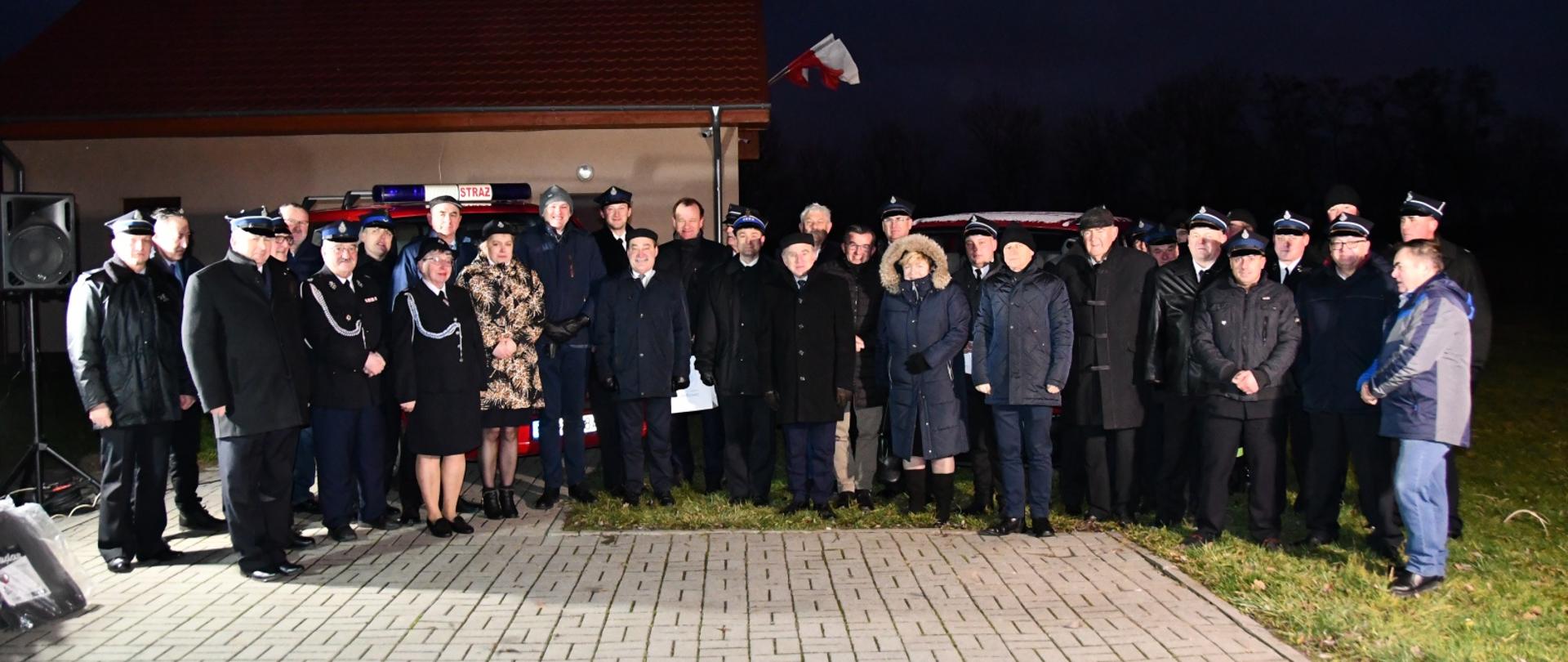 Grupa strażaków i osób cywilnych na tle samochodów pożarniczych