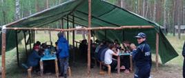 na zdjęciu namiot w lesie na polanie pod którym siedzą przy stołach uczestnicy obozu do nich idzie funkcjonariuszka polucji 