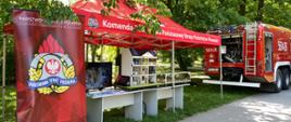 Stoisko straży pożarnej na festynie z okazji dnia dziecka. Symulator zagrożeń w domu jednorodzinnym oraz woda-las, a także wóz bojowy straży pożarnej.
