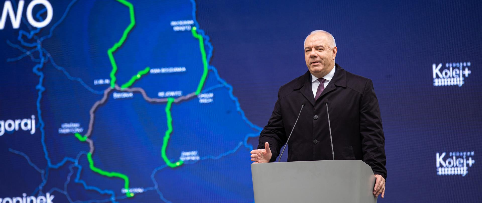 Wicepremier Jacek Sasin stoi przy mównicy. W tle ścianka z mapą Polski i logotypami Kolej+