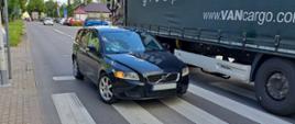 Czarny samochód osobowy, kombi, marki volvo, który stoi na jezdni, na pasach, na których doszło do potrącenia. Po prawej stronie widoczny jest samochód ciężarowy, który mija samochód osobowy.