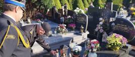 Widok z boku. Od lewej widoczny stojący przed grobem strażak. Obok drugi strażak pochyla się i stawia na mogile zapalony znicz. W tle inne groby.