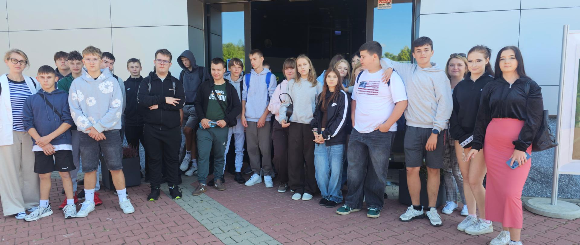 Grupa uczniów i opiekunów stoi przed wejściem do nowoczesnego budynku podczas wspólnego wyjścia edukacyjnego.