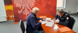 Na zdjęciu widzimy dwóch mężczyzn siedzących przy stoliku i podpisujących dokumenty. Stolik koloru brązowego. Mężczyzna z prawej strony kadru to strażak, ubrany w mundur galowy kolor czarnego. Mężczyzna z lewej to cywil ubrany w granatową bluzę oraz oraz spodnie. Mężczyźni w lewych dłoniach trzymają dokumenty. W tle widzimy czerwony baner koloru czerwonego z godłem polski oraz napisem Państwowa Straż Pożarna. 