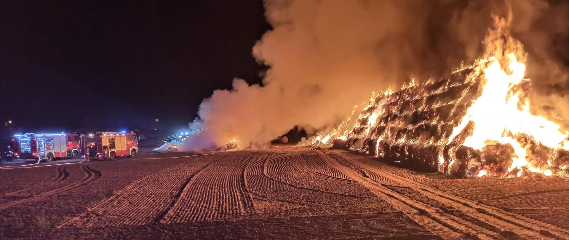 Pora nocna. Pali się stóg słomy w całej objętości. Widać ogień i duże zadymienie. Obok stogu stoją wozy pożarnicze z włączonymi światłami błyskowymi. Trwa akcja gaśnicza.