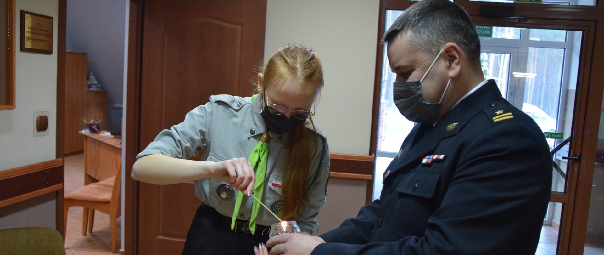 Harcerka przekazuje przy pomocy cienkiego drewnianego patyczka ogień do świecy w szkle, którą trzyma strażak w umundurowaniu galowym.