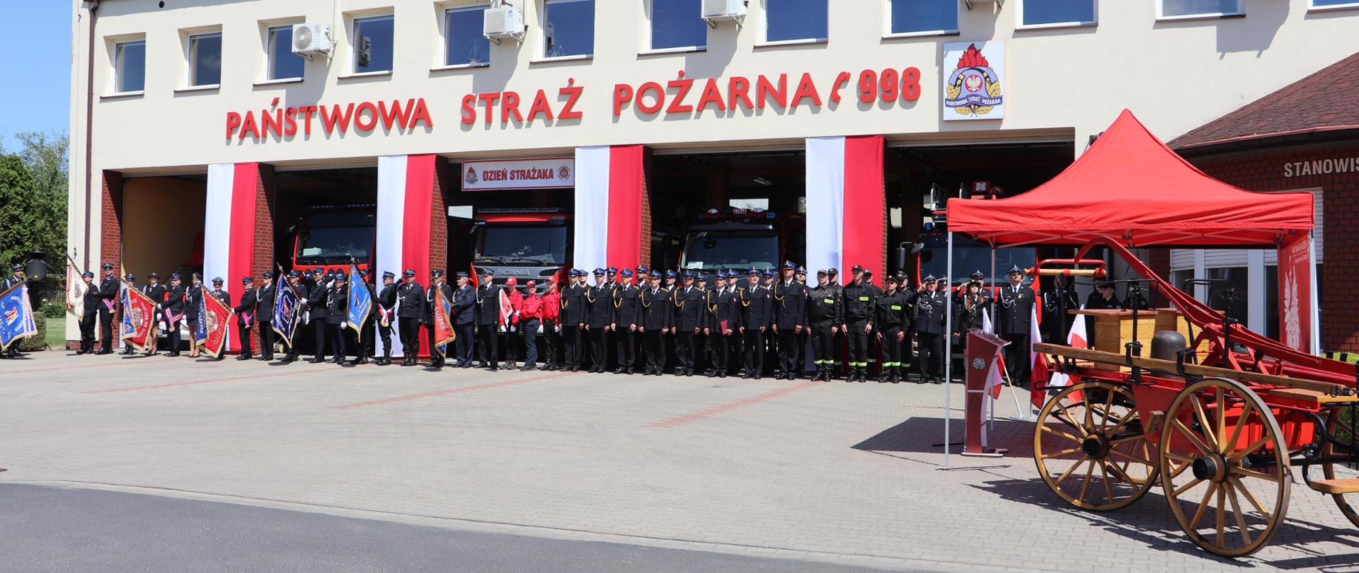 Pododdziały pocztów sztandarowych oraz strażaków PSP, OSP oraz MDP podczas Powiatowych Obchodów Dnia Strażaka w Środzie Wielkopolskiej na tle budynku strażnicy.