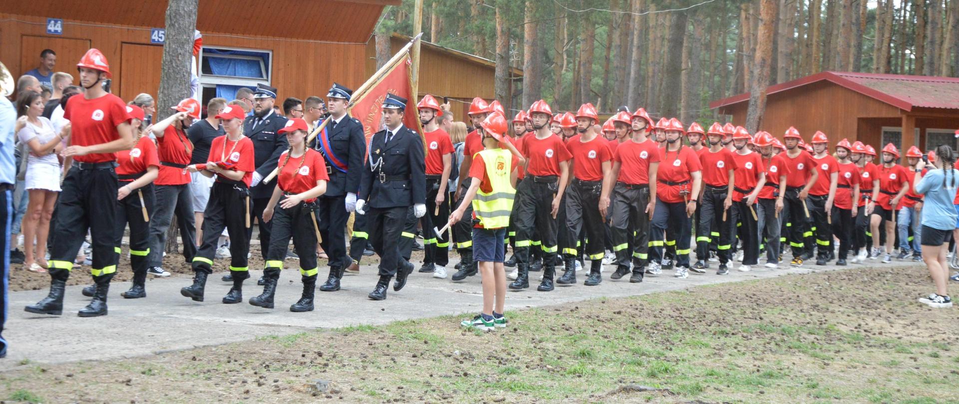 Teren ośrodka wypoczynkowego. Betonową drogą pomiędzy drewnianymi budynkami na plac apelowy maszeruja kompanie obozowiczów . Na sobie mają czerwone koszulki i czerwone hełmy. 