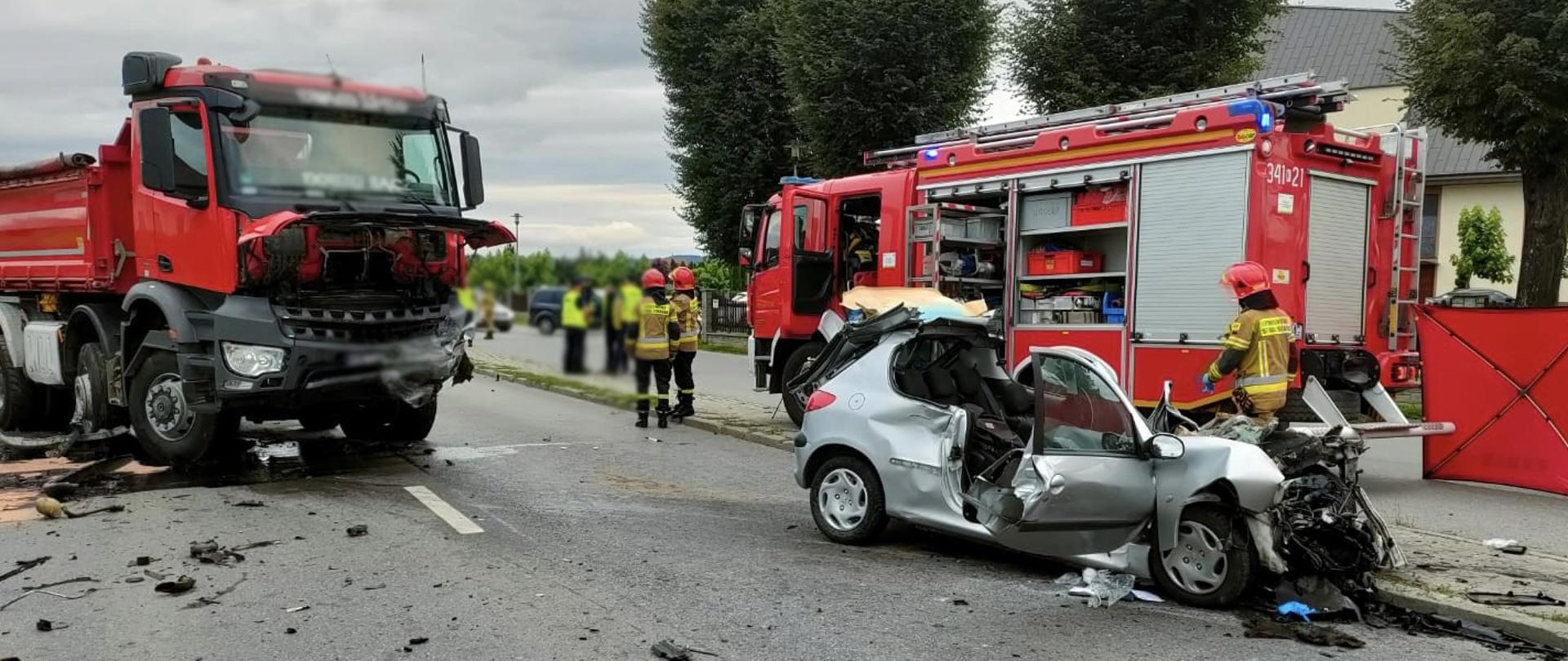 Rozbite samochody ciężarowy i osobowy na drodze a obok samochód pożarniczy i strażacy