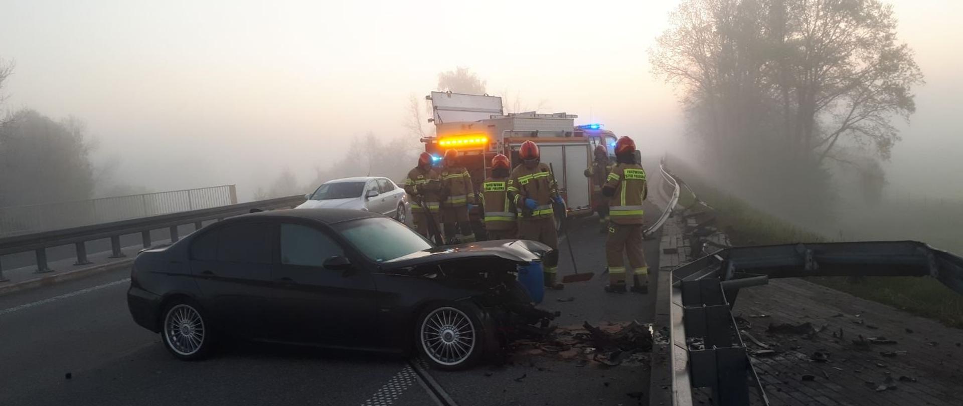 Poranna mgła nad drogą. Centralnie na zdjęciu rozbite czarne auto w poprzek drogi, uszkodzone barierki z prawej strony. Z nim strażacy i samochód strażacki.